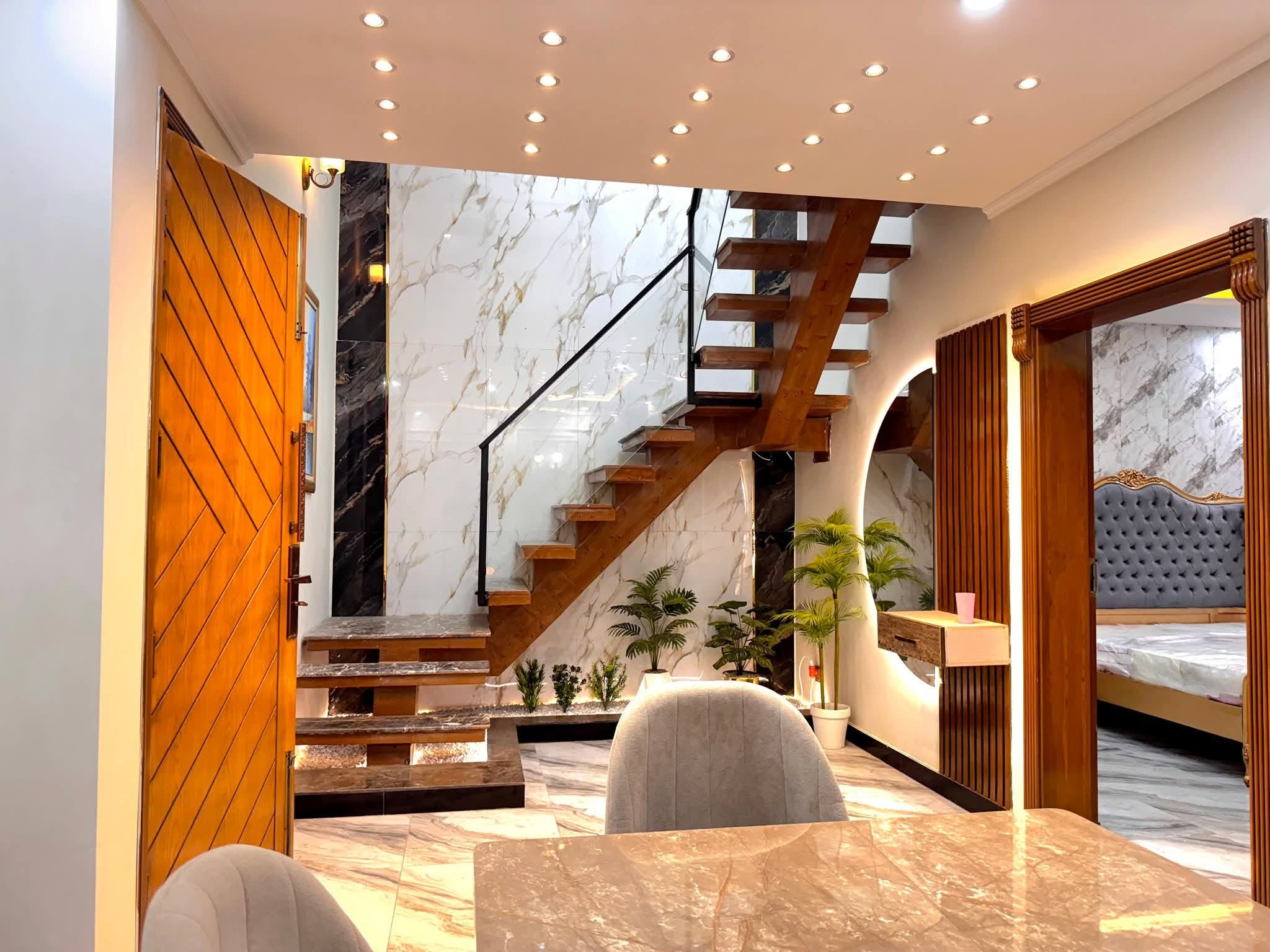 Inside a finished MK Builders home — the wooden front door opens onto a marble-clad entrance hall with a floating timber staircase, glass balustrade, and recessed ceiling lights.